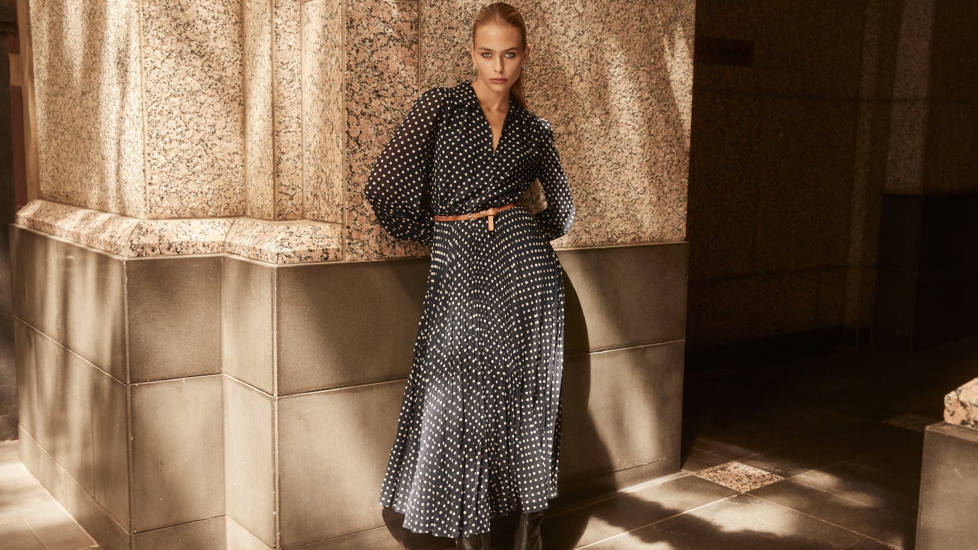 model wearing a polka dot pleated midi dress standing against a large stone wall with dappled sunlight