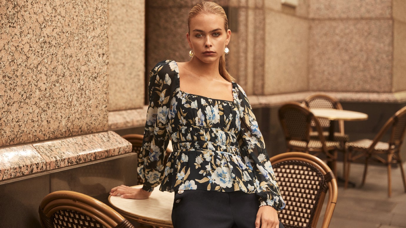 model posing in front of brown building with french-style bistro outdoor chairs wearing a dark blue floral long sleeve blouse