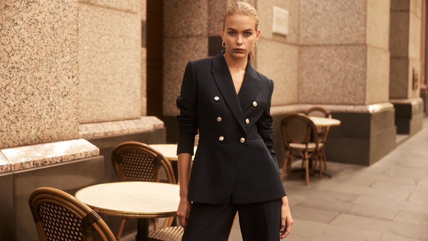 model standing outside a large granite looking building with french-style bistro chairs on the footpath wearing a navy suit with gold buttons