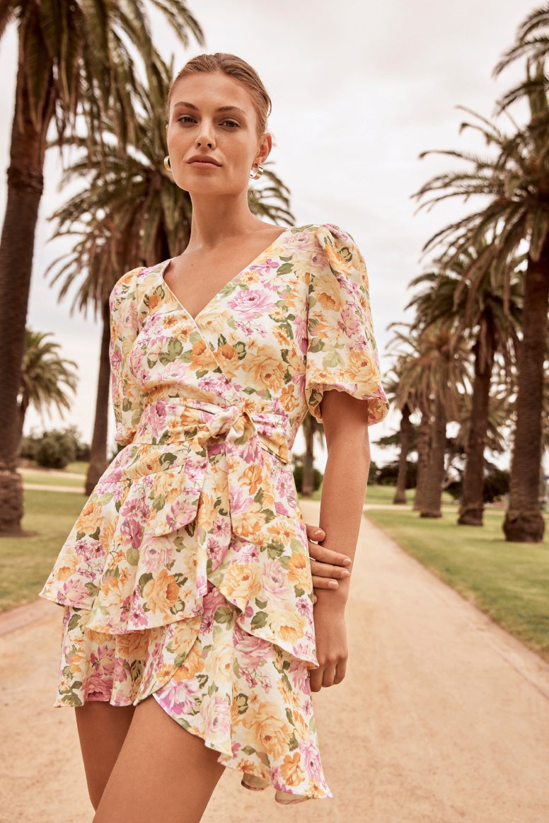 model in pink and yellow floral print mini dress with tiered design standing on a sandy path with pam trees in the background