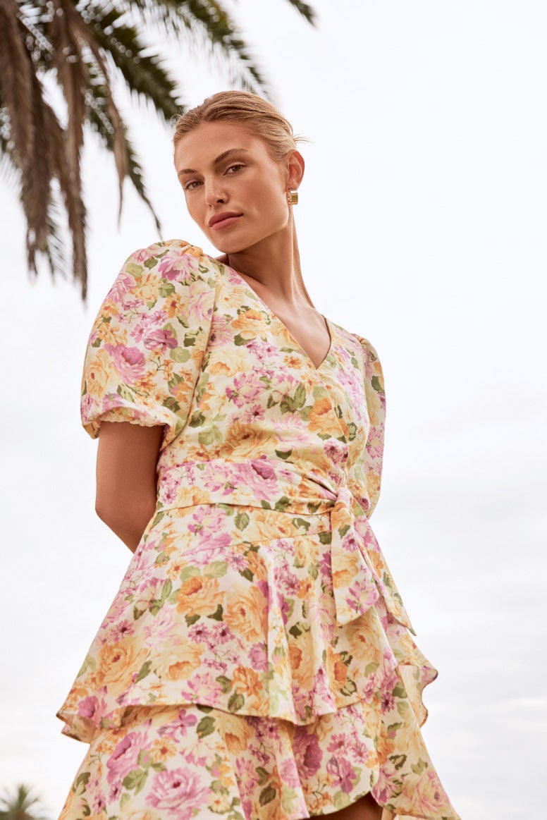 model in pink and yellow floral mini dress with puff sleeves and tiered design with palm tress in the background