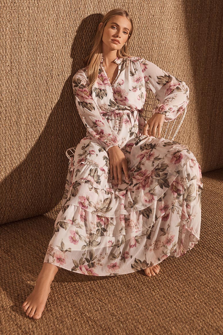 a blonde barefoot model sits and poses on a chair wearing a long sleeve floor length pink and cream floral dress, the studio setting has warm golden hessian or jute looking wall and floor coverings