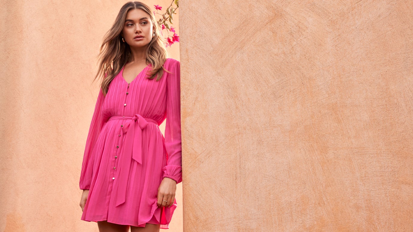 model in a pleated pink mini dress with long sleeves standing against a terracotta wall