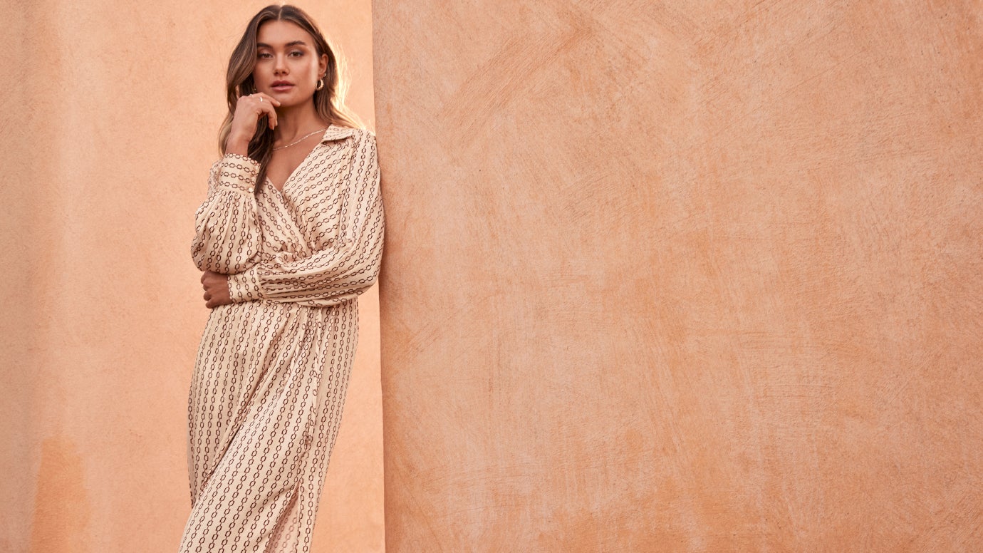 model in a cream chain link print shirt dress in satin crossing her arms standing in front of a terracotta wall