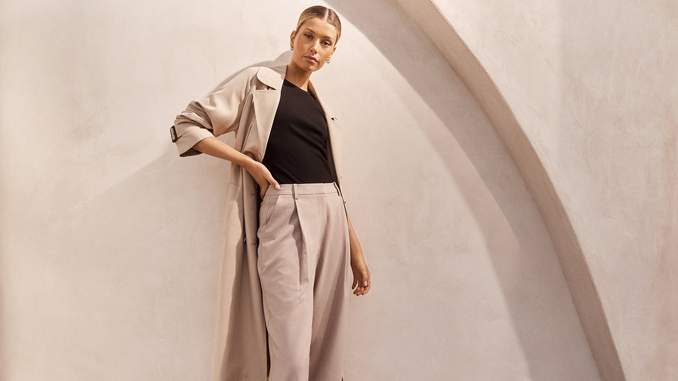 model wearing a soft stone coloured trench and matching wide-leg pants with a plain black top, she is posing with one hand on her hip