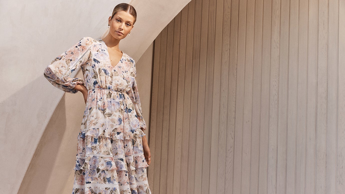 a model with blonde hair slicked back wearing a white tiered midi dress with a blue and lilac floral print posing in front of a concrete rendered arch and wooden paling