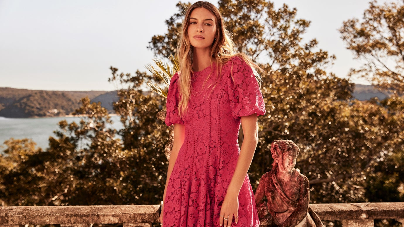 model in deep red mini dress with puff sleeves and all-over detailed rose applique standing in on a balcony with mediterranean looking background including the sea