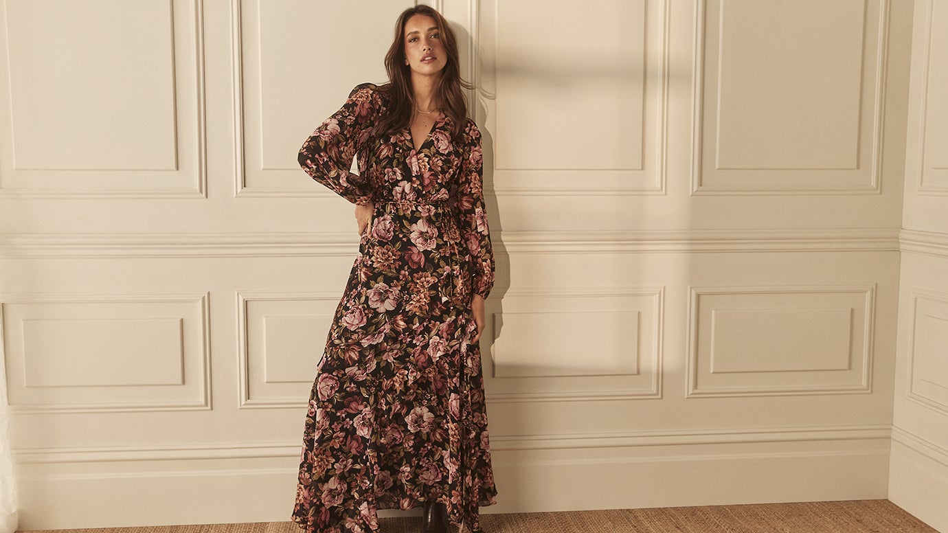 a brunette model wearing a long flowing tiered dress with long sleeves in bronze black and plum floral print standing against an elegant cream coloured panelled wall