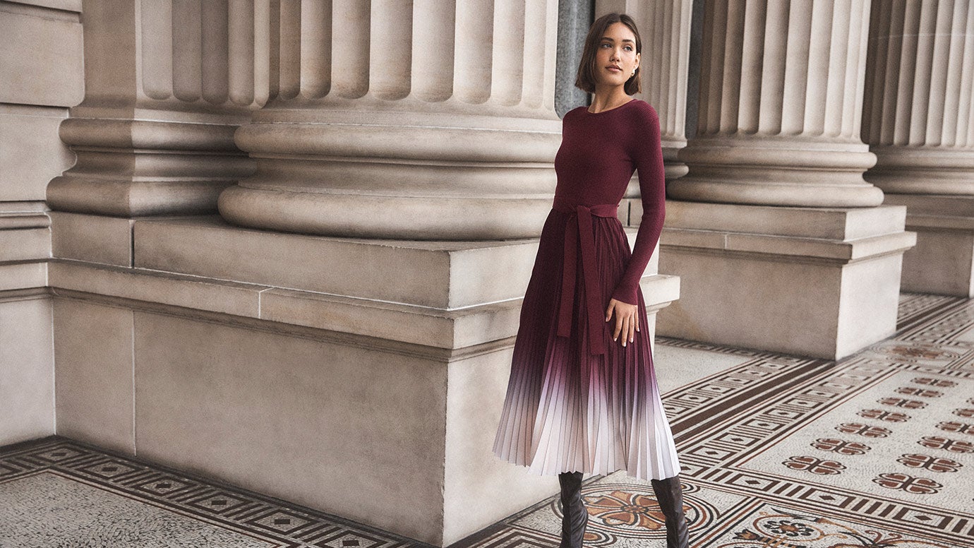 model with brown bob wearing a dress that looks like a long sleeve knit top in plum with a pleated plum and white ombe skirt and tall brown boots, she poses near enormous old pillars and a patterned paved floor