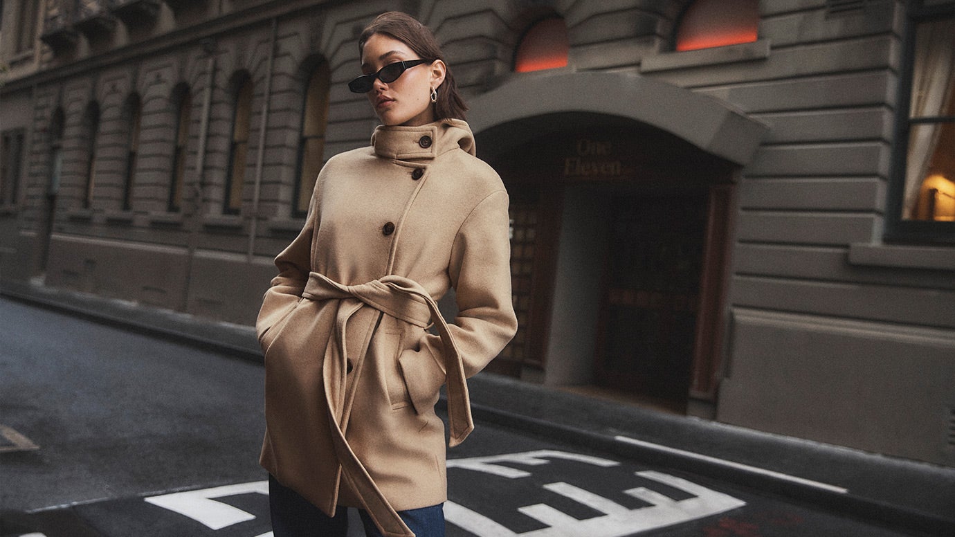 model with brown bob wearing dark rectangular sunglasses wearing a camel coloured hooded belted jacket standing on a deserted city side street