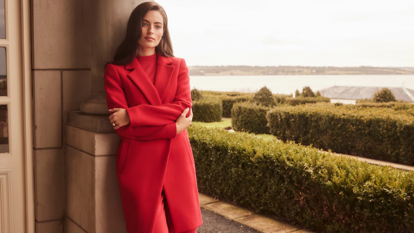 model leaning back against a large column wearing a bright red coat and matching pants with a classic regal looking garden with hedges in the background