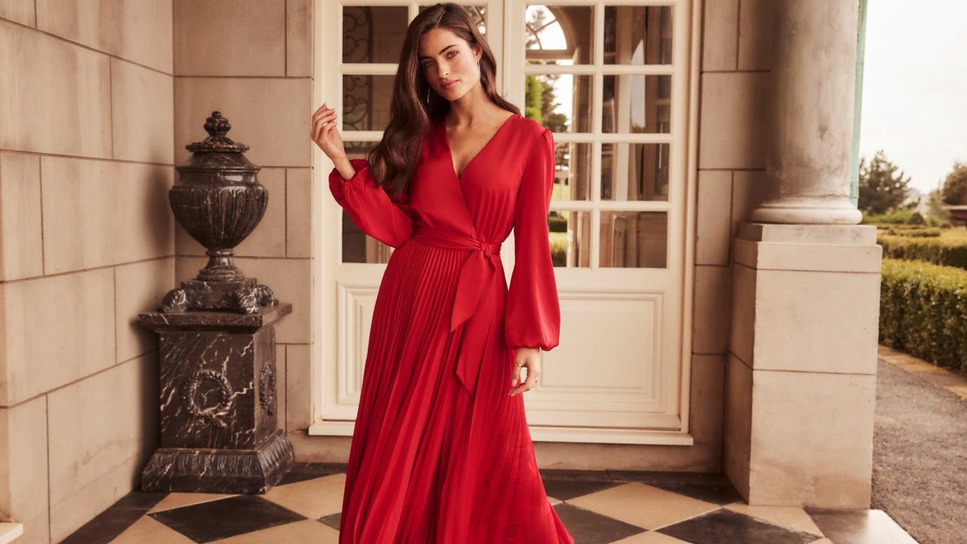 model wearing a pleated wrap midi dress in deep red standing in front of french doors of a grand sandstone building