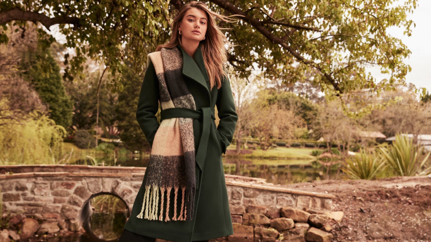 model standing in a leafy countryside area wearing a dark green coat and a cream checked scarf with her hands in her pockets