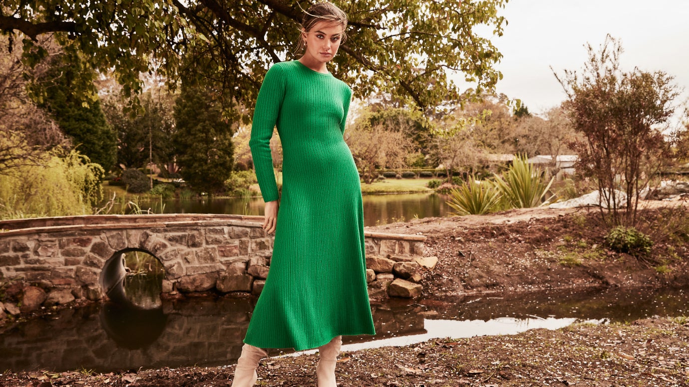 model wearing a ribbed knit dress with an a-line skirt in bright green with cream boots with a pond behind her and a quaint stone bridge