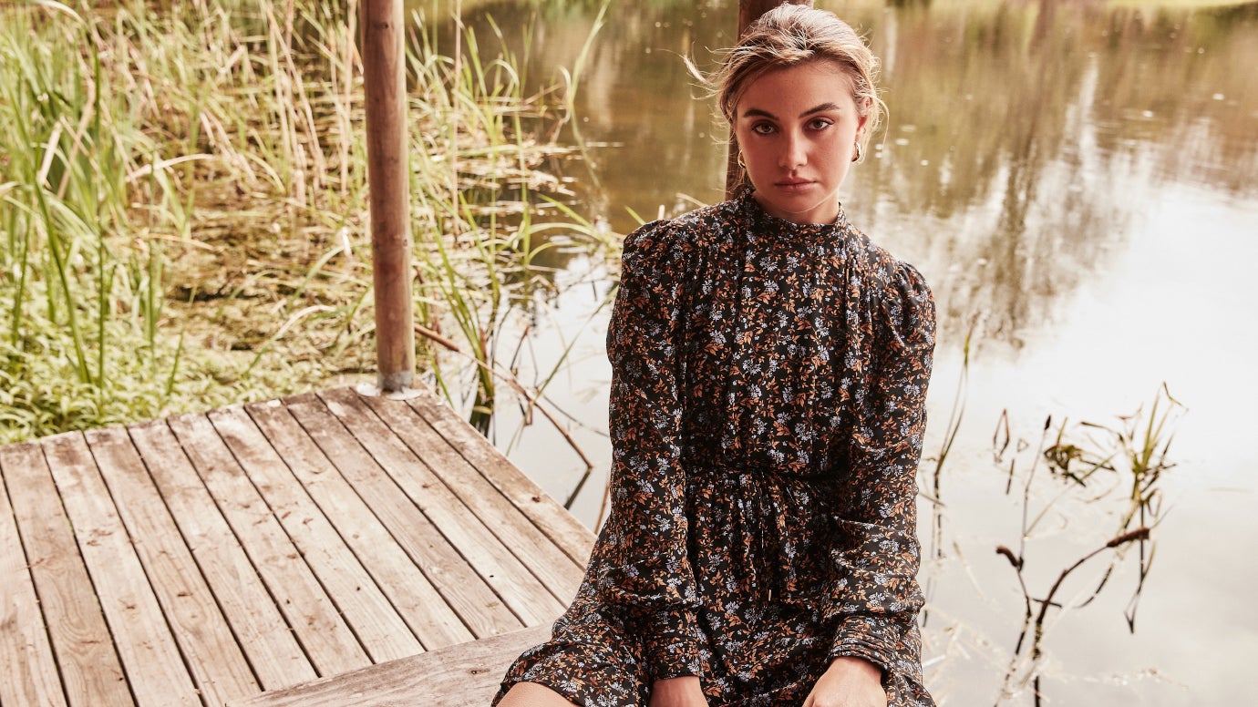 model sitting on dock near a tranquil pond wearing a high neck mini dress with delicate floral print in shades of deep brown and blue