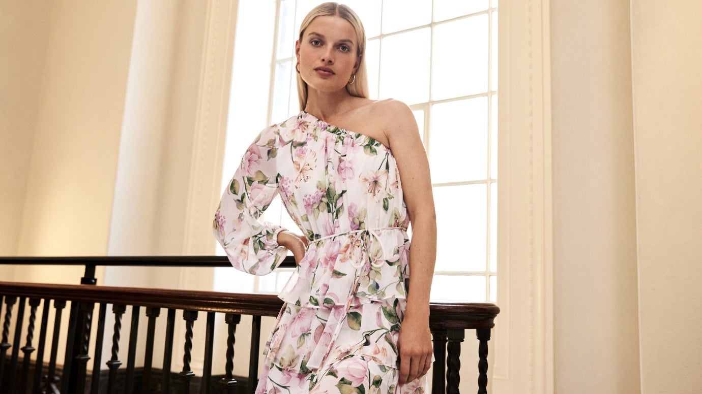 model standing against vintage wooden staircase railing with one hand on hip wearing a one sleeve mini dress in a pink green and white floral print