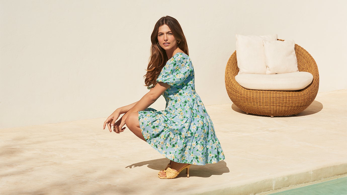model in blue mini dress with white and yellow floral print and thatched style leather mules crouching by a resort style pool 