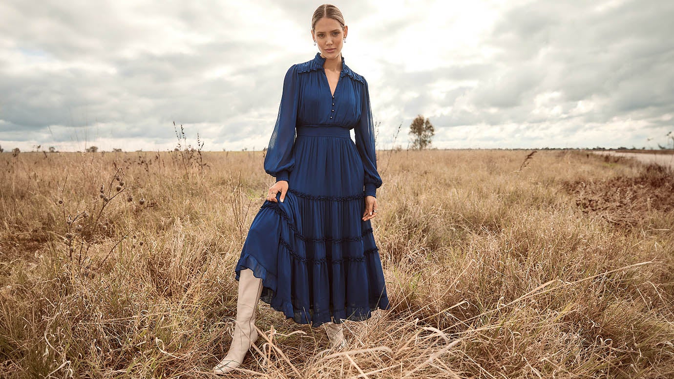 a model wearing a long flowing dress in deep blue with sheer long sleeves and cream knee high boots, she is standing in an empty field with long wheat and under an expansive cloudy sky