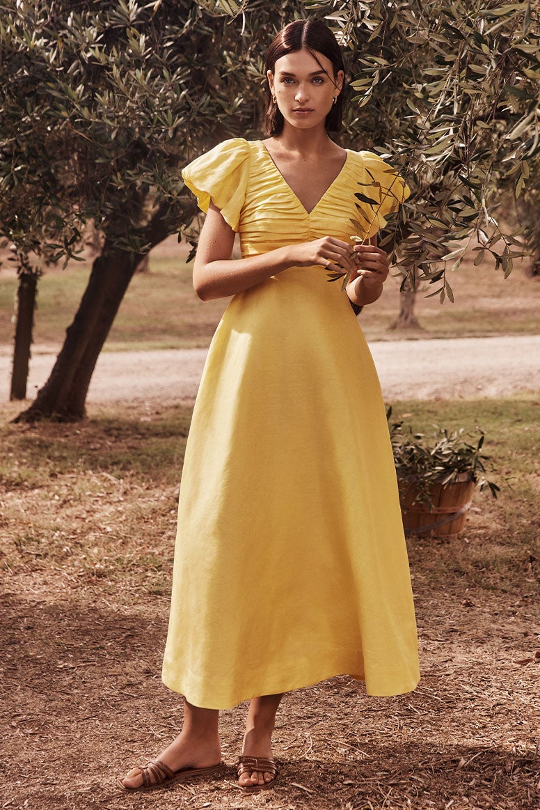 model with brown bob wearing an airy yellow linen dress with a v neck, ruching on the bust, and statement ruffle sleeves and brown slides, she is posing in an olive grove, she is holding onto a small branch 