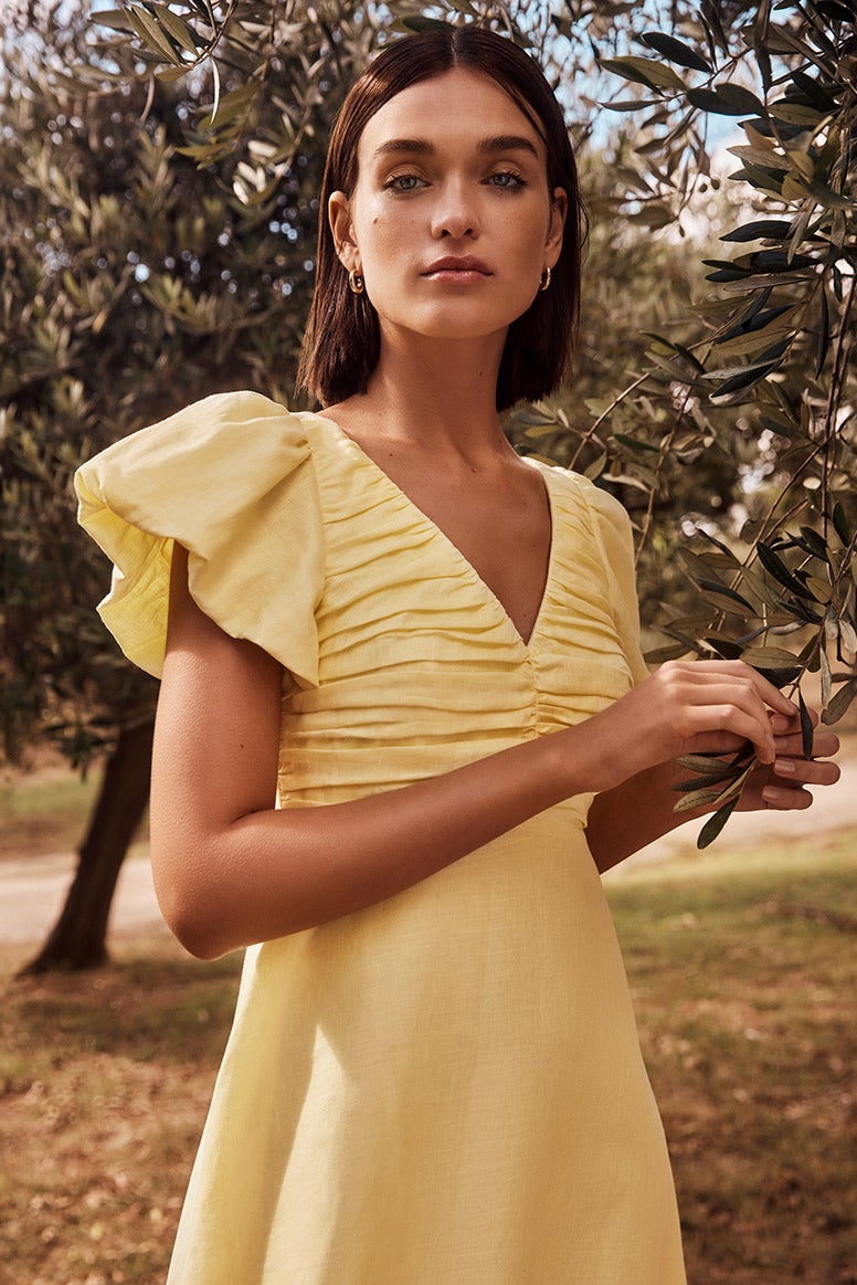 model with a brunette bob wearing an airy yellow linen dress with statement ruffle sleeves posing in an olive grove, she is holding onto a small twig from our olive tree, the photo focuses on her face and upper body