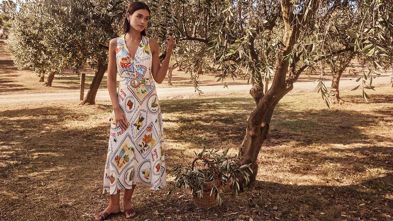 model with brown bob wearing a sleeveless dress with a belted waist with a straw buckle, the dress has a Mediterranean themed postcard print, the model poses in a quaint olive grove near a woven basket filled with olive branches