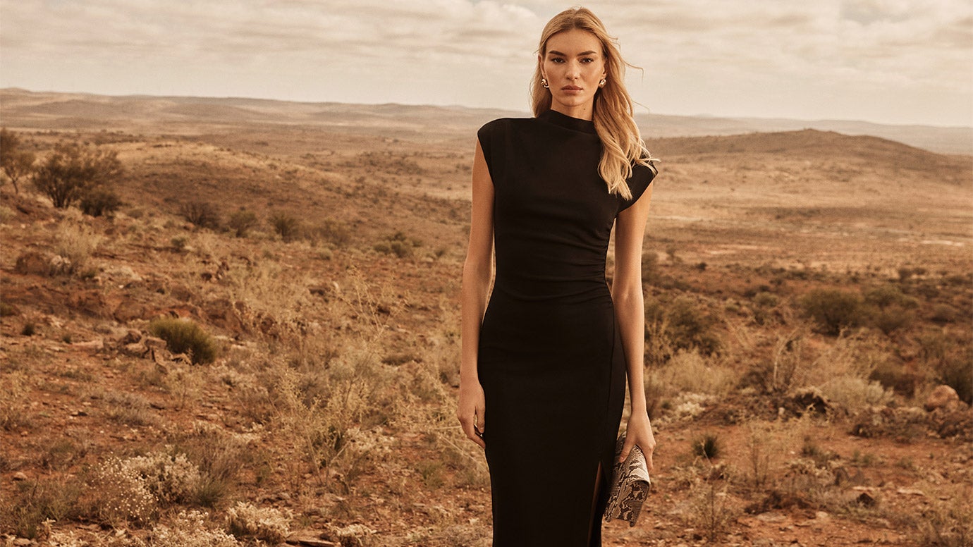 blonde model wearing a classic black cocktail dress with a high neck poses in a remote landscape that looks hazy and dusty, she holds a snakeskin clutch in one hand by her side