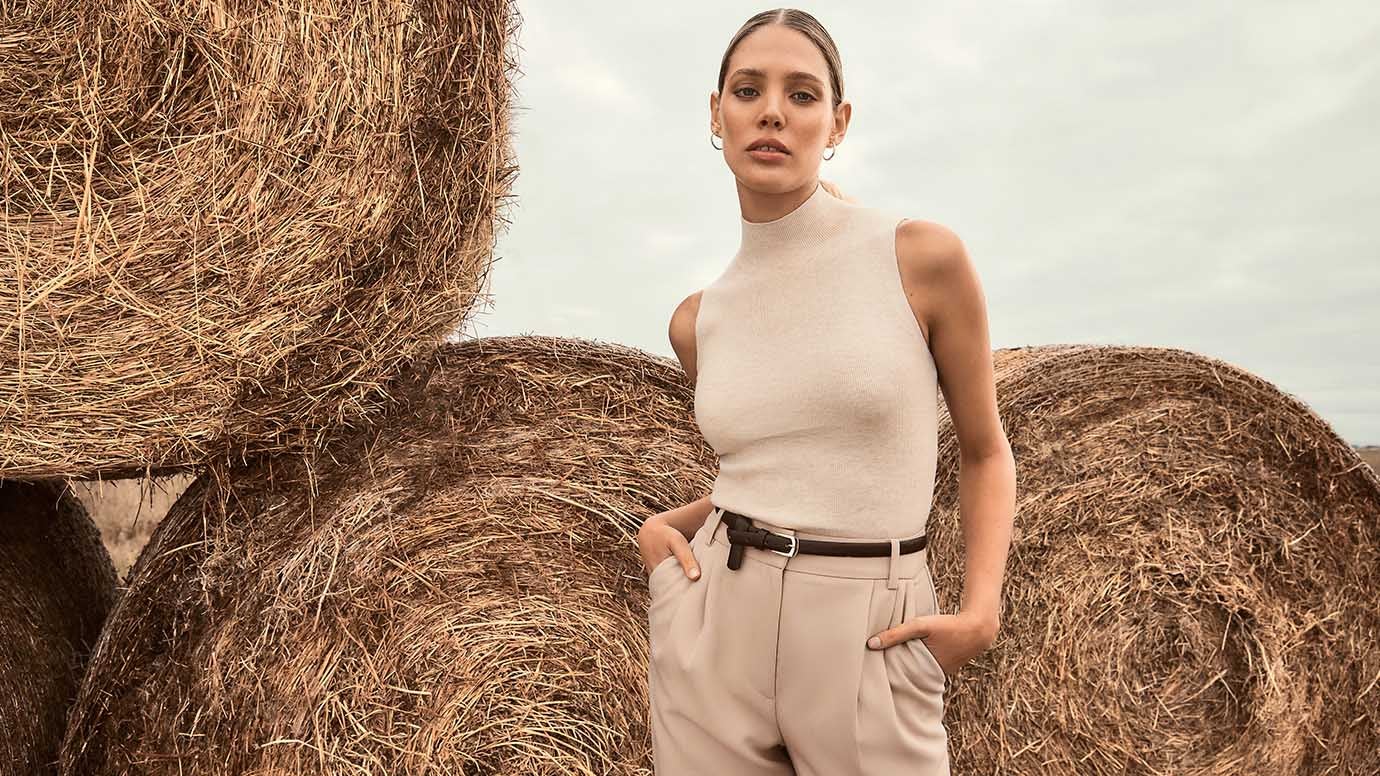 a model wearing a taupe high neck sleeveless knit top with beige tailored pants and a skinny belt poses in front of large rolled hay bales with her hands in her pockets