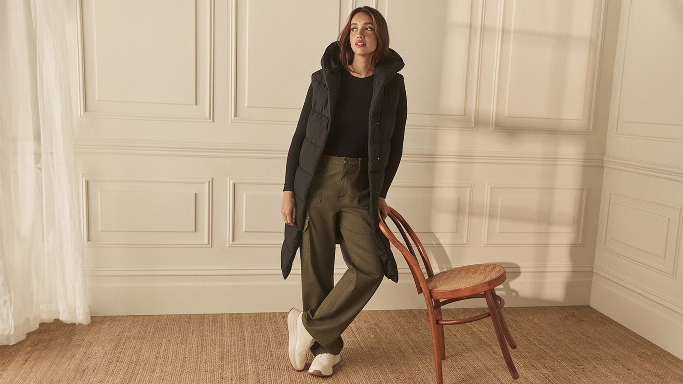 model wearing a black puffer longline vest with a hood and a black basic top underneath with khaki green cargo pants and white sneakers poses before a cream coloured panelled wall leaning against an elegant wooden chair