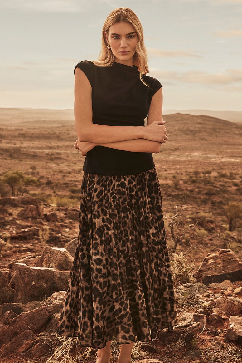 a blonde model poses with her arms crossed in front of her wearing a black draped top with a high neck and short sleeves and leopard print skirt, the landscape behind her looks remote and dusty with mountains