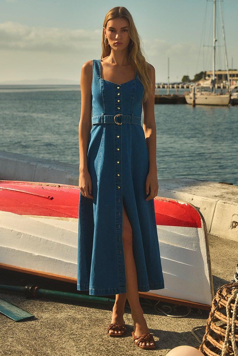 model wearing a long denim dress with silver buttons down the front and wide tank top style straps stands in front of an upturned rowing boat, boats and blue water is seen in the background