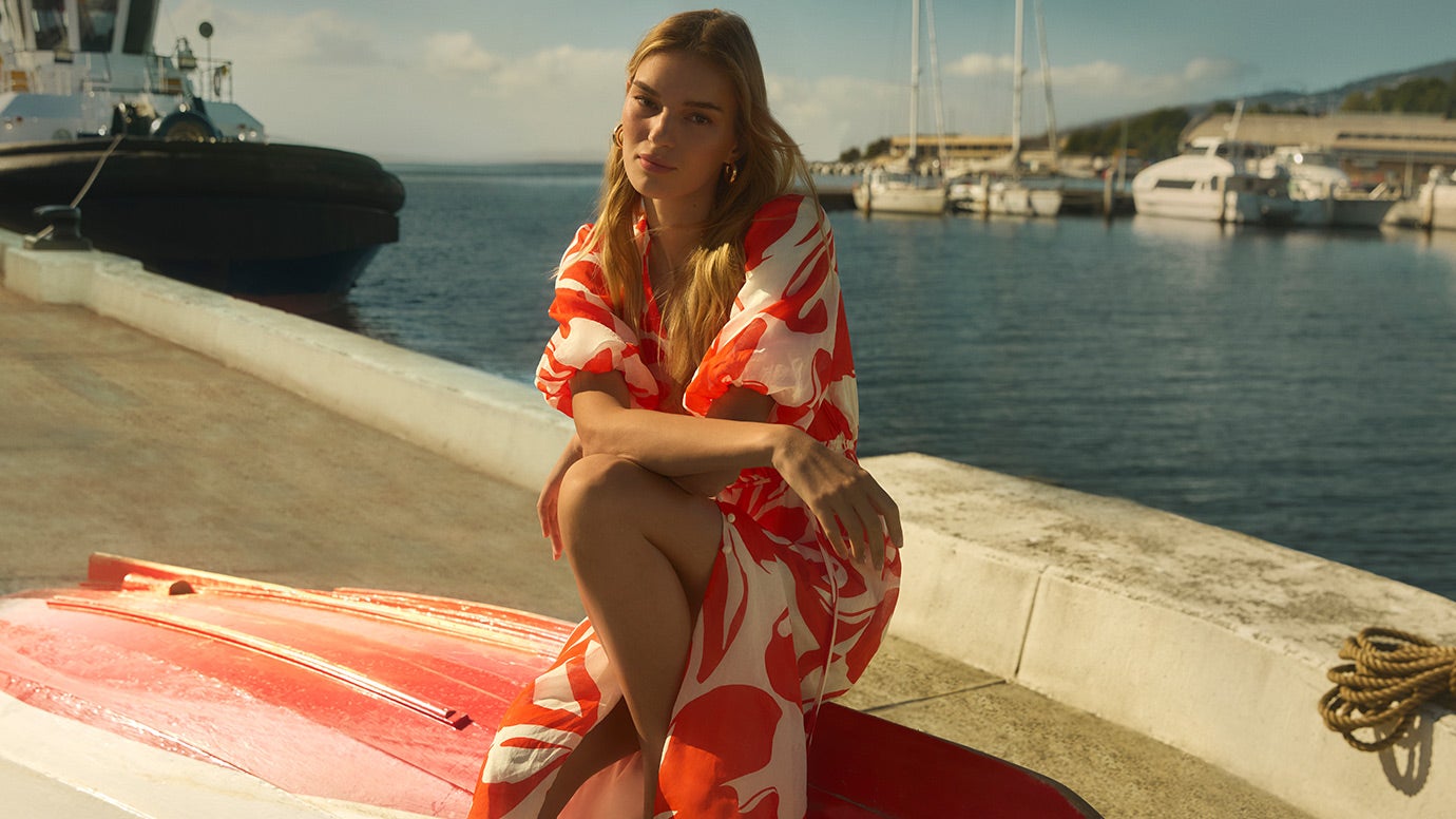 a blonde model crouches on an upturned red and white row boat, she is dressed in a red and white sundress with puff sleeves, the water and big boats are seen in the background