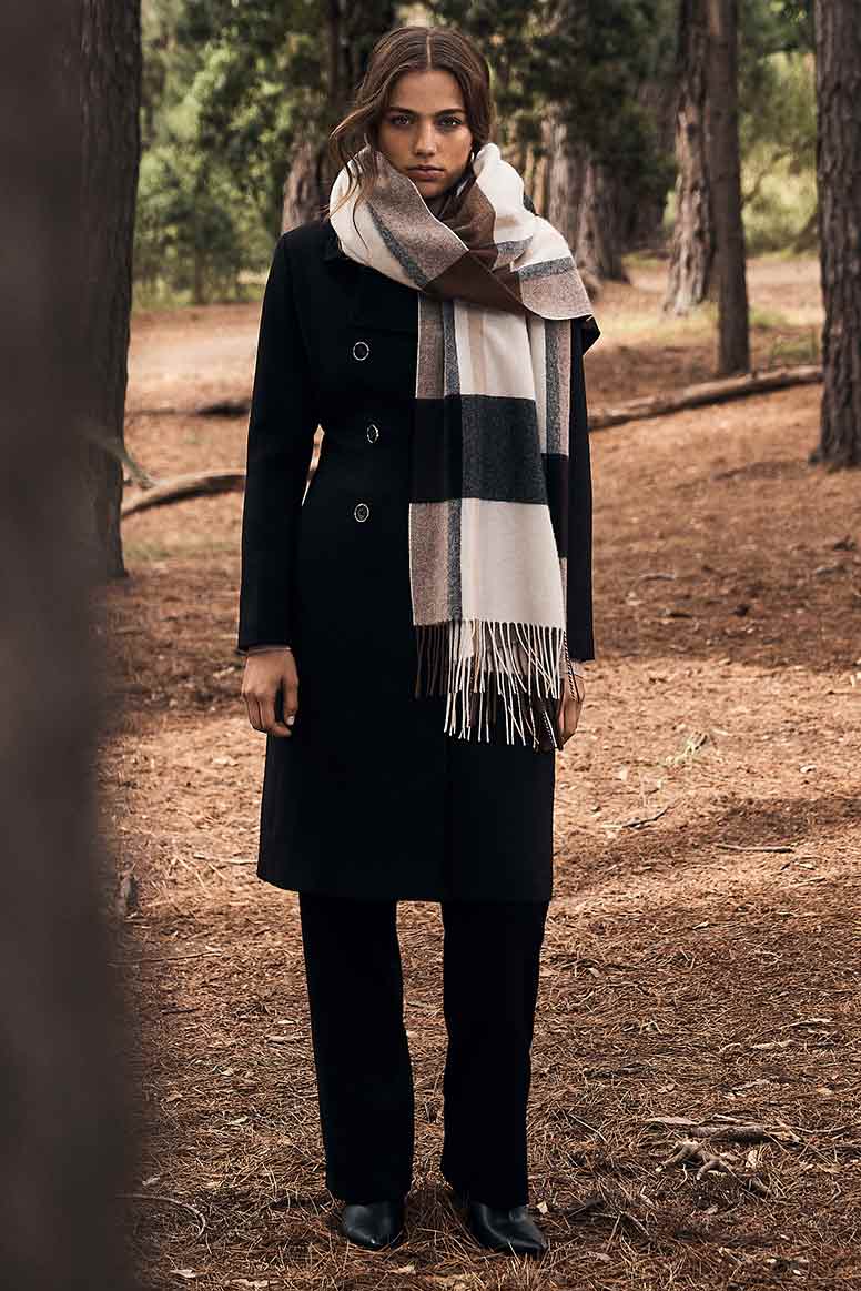 a model wearing a long black coat and black pants and a big check cozy looking scarf in tones of brown and cream stands in a forest, a tree trunk is in the fore ground