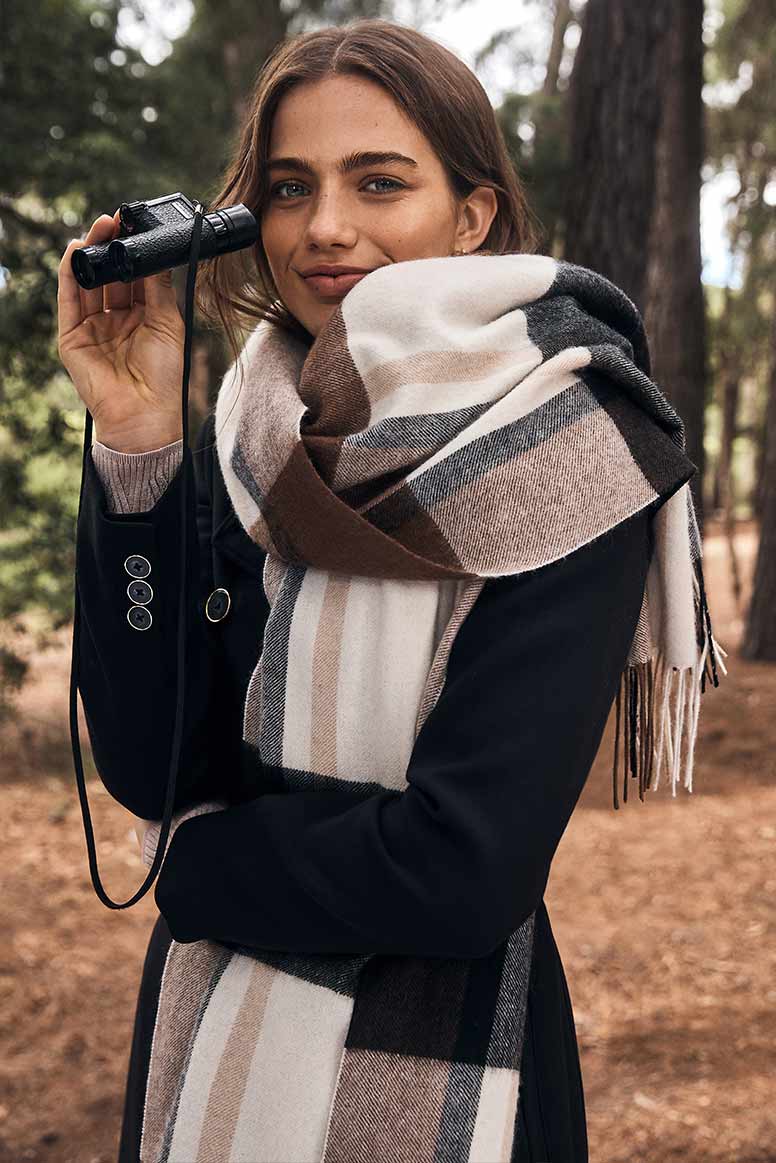 a model poses in a forest with bonculars in one hand, she is wearing a black coat and large cozy scarf in a check print, the scarf is different shades of cream beige and brown