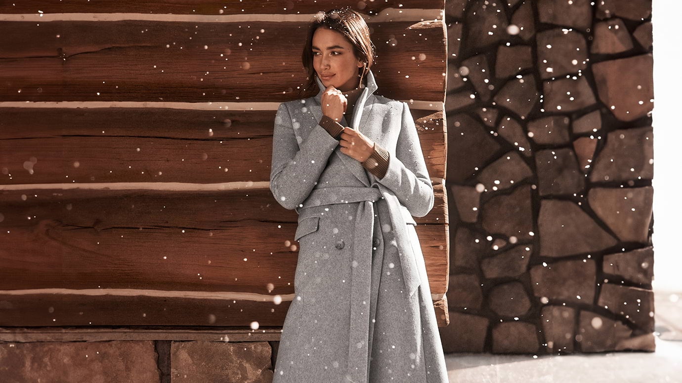 a brunette model posing in the snow just outside a log cabin, she is wearing a long grey coat with a belt and a brown knit top, she is holding the coat together at the bust