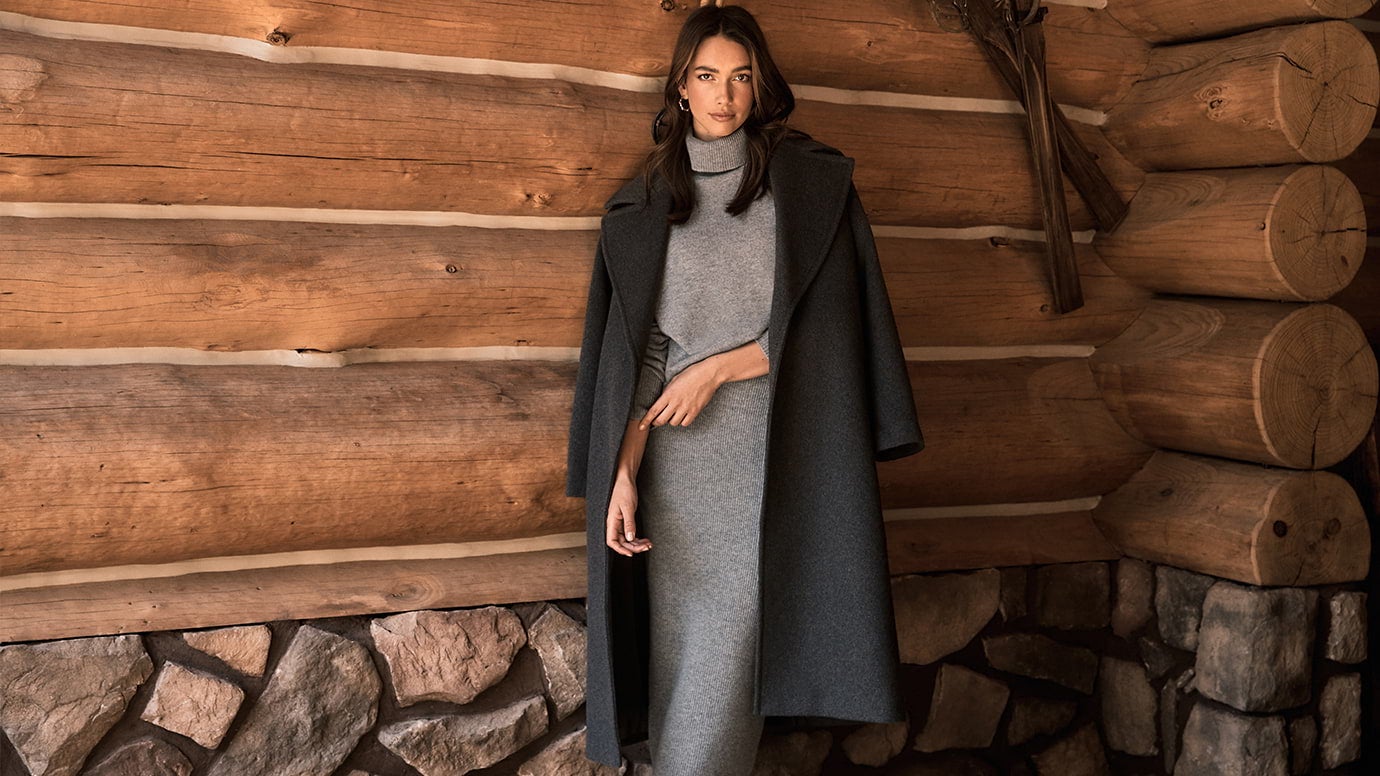 a brunette model posing in frontof a log cabin wall wearing a long charcoal coat with a lighter grey knit set on underneath, she is crossing her arm against herself