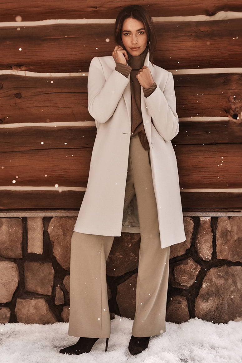 a brunette model posing against the wall of a log cabin while snowflakes fall around her, she is wearing a brown sweater, camel pants, heel boots and a cream coat
