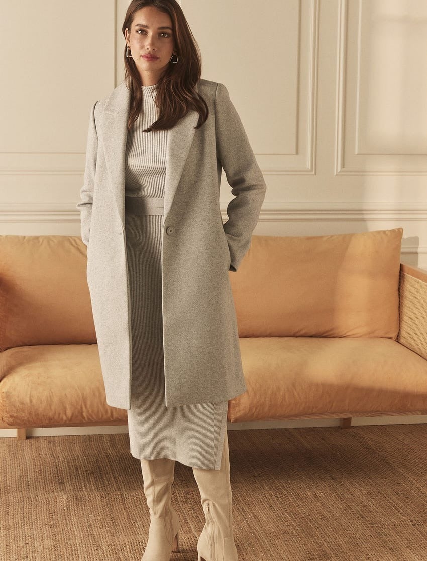 model wearing a light coat with structured shoulders in light grey posing in front of a tan leather sofa, she has a similar coloured knit dress on underneath and cream boots on