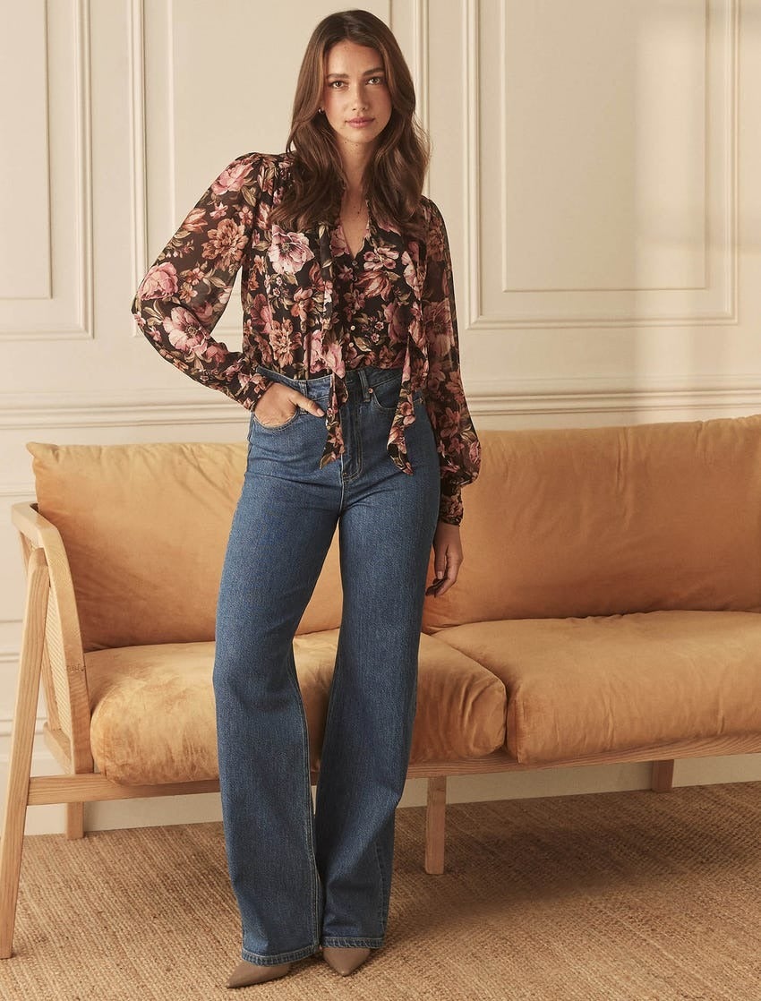 model wearing a floral blouse in tones of plum and camel with mid blue straight leg jeans and pointed pumps poses in front of a tan leather sofa