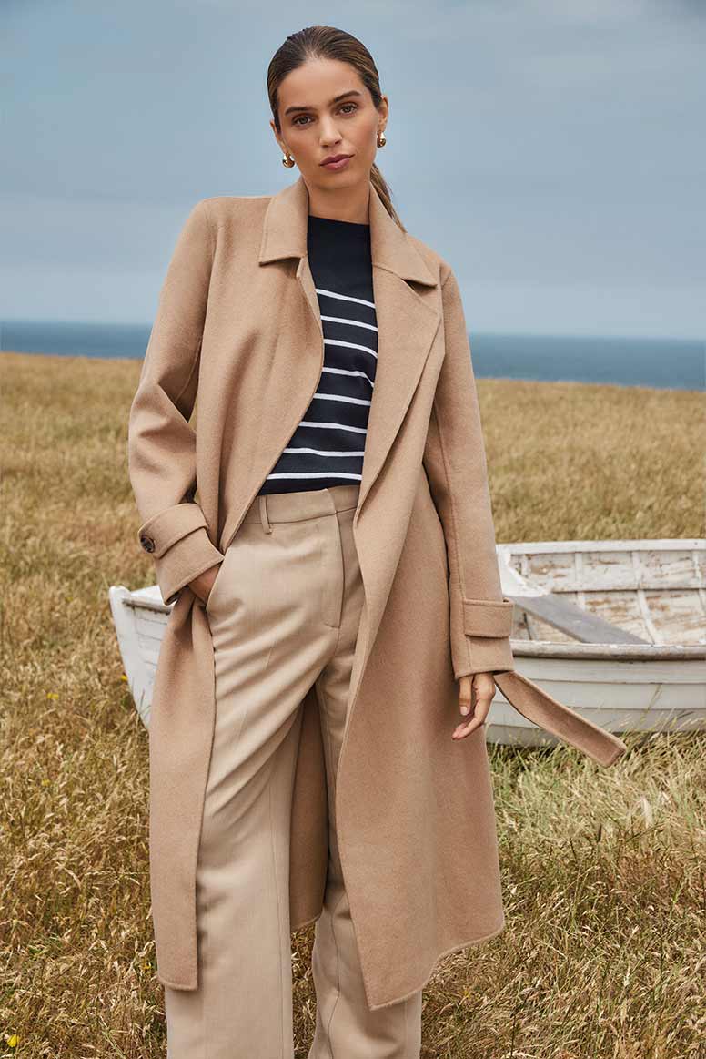 a model wears a camel coat, a striped navy sweater and beige pants, she stands on sunbleached grass on a cliff, a whitewashed old boat, the sea, and blue sky is behind her