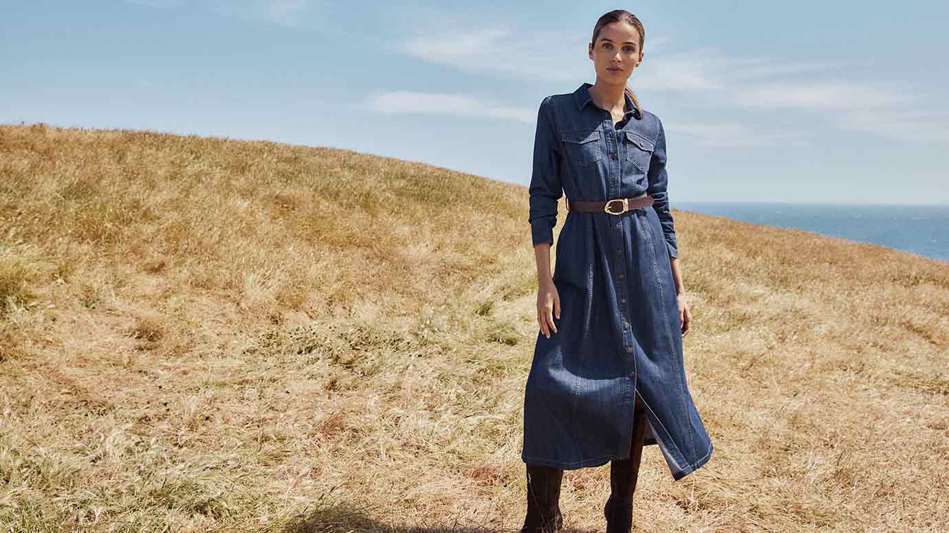 model wears a deep blue denim shirt dress with a dark brown belt and keen high boots, she stands on a sunbleached grass covered hill the blue sky is see behind her