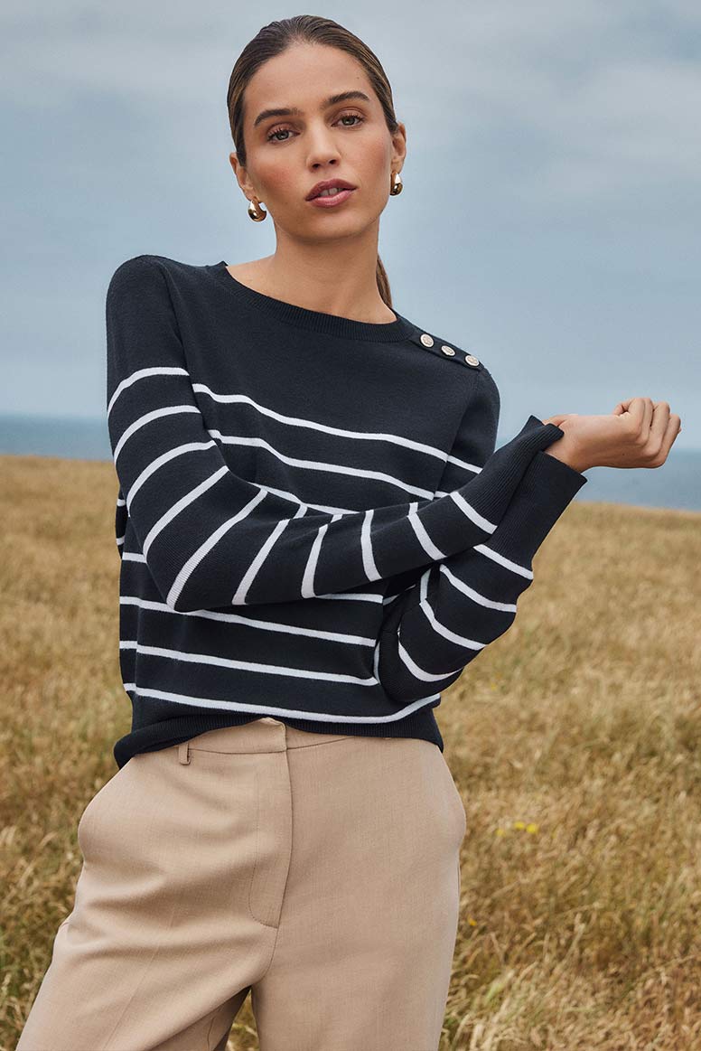 a brunette model in a navy sweater with white stripes and stone coloured pants, she is posing against a cloudy sky and you can see sun bleached grass in the background