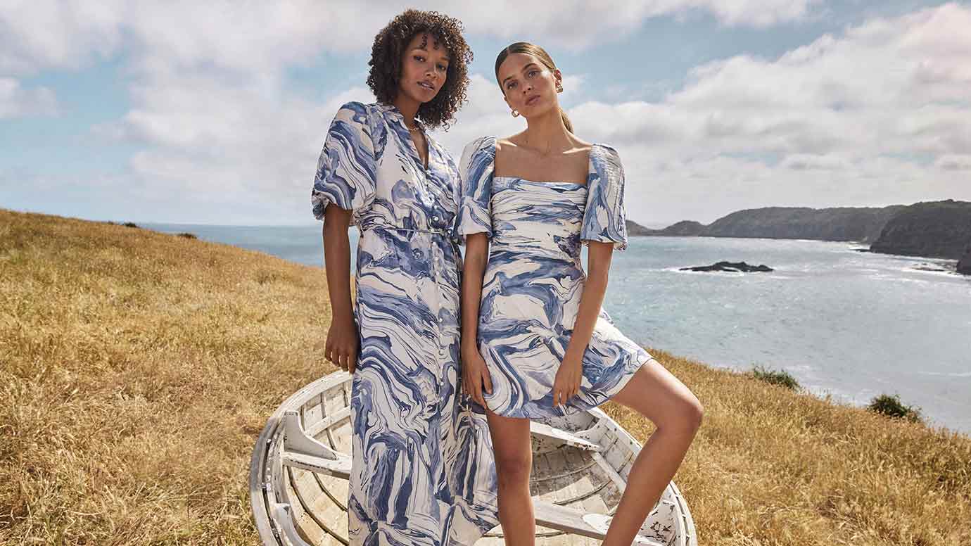 two models pose together in blue and white marble print dresses atop a cliff overlooking a turquoise ocean, the black model has soft curly hair and is in the longer dress while the brunette model wears the mini version