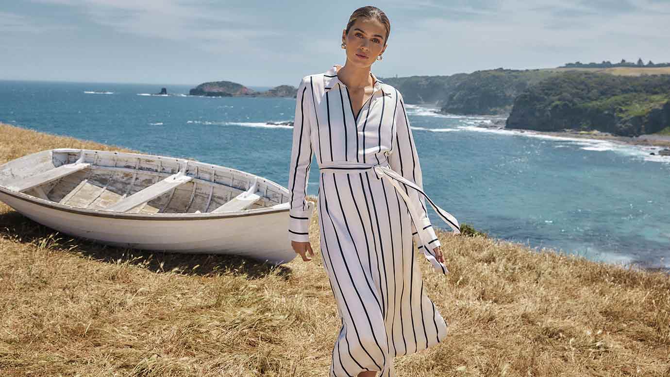 brunette model wearing a satin shirt dress in cream with black vertical stripes, she is standing atop a cliff covered in sun bleached grass, turquoise ocean is seen below in the background