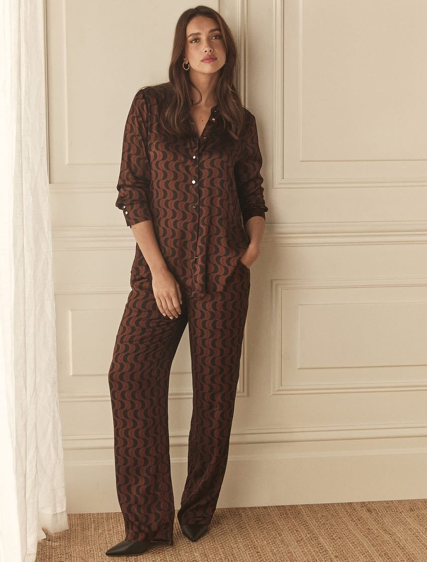 brunette model wearing a satin shirt and matching relaxed pants in a deep choc brown wavy line geo print, she poses in front of a wainscoted wall