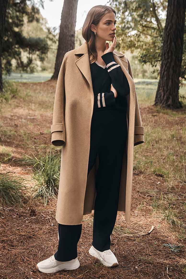 a model poses looking to the right in a forest location wearing a black knit top and matching pants with camel coloured stripe detailing and a camel coat and white sneakers