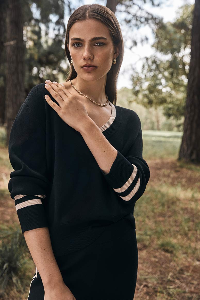 model poses in a forest setting, she is wearing a black knit sweater with camel coloured stripe detail and black pants 