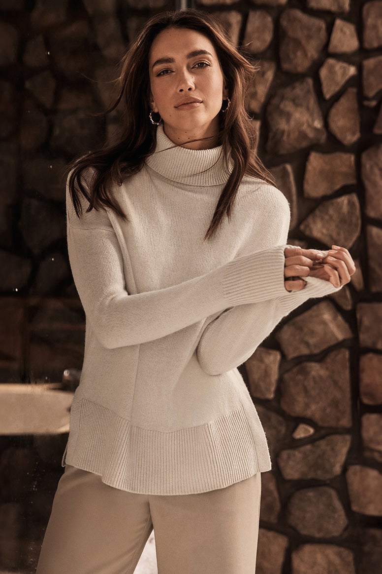 a brunette model poses in front of a rock wall or column wearing camel pants and a cream roll neck jumper, you can see a few small snowflakes in the air behind her