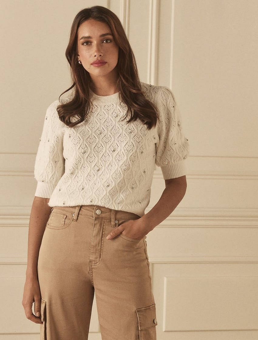 a brunette model wearing a short sleeve cream sweater with diamond shaped pointelle detailing and small clear diamante jewels attached, and camel tailored cargo pants poses with her hand in her pocket