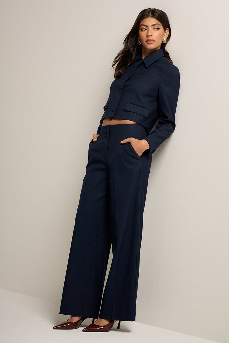 model with dark hair leans against white studio wall, she is wearing pointed toe heels and a matching tailored set in navy, the jacket is slightly cropped, the pants are relaxed and you can see a little bit of her midriff