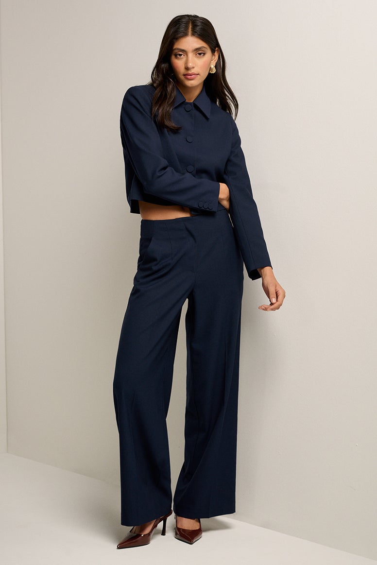 model with dark hair stands in a white studio setting she is wearing pointed toe heels and a matching tailored set in navy, the jacket is slightly cropped, the pants are relaxed and you can see a little bit of her midriff